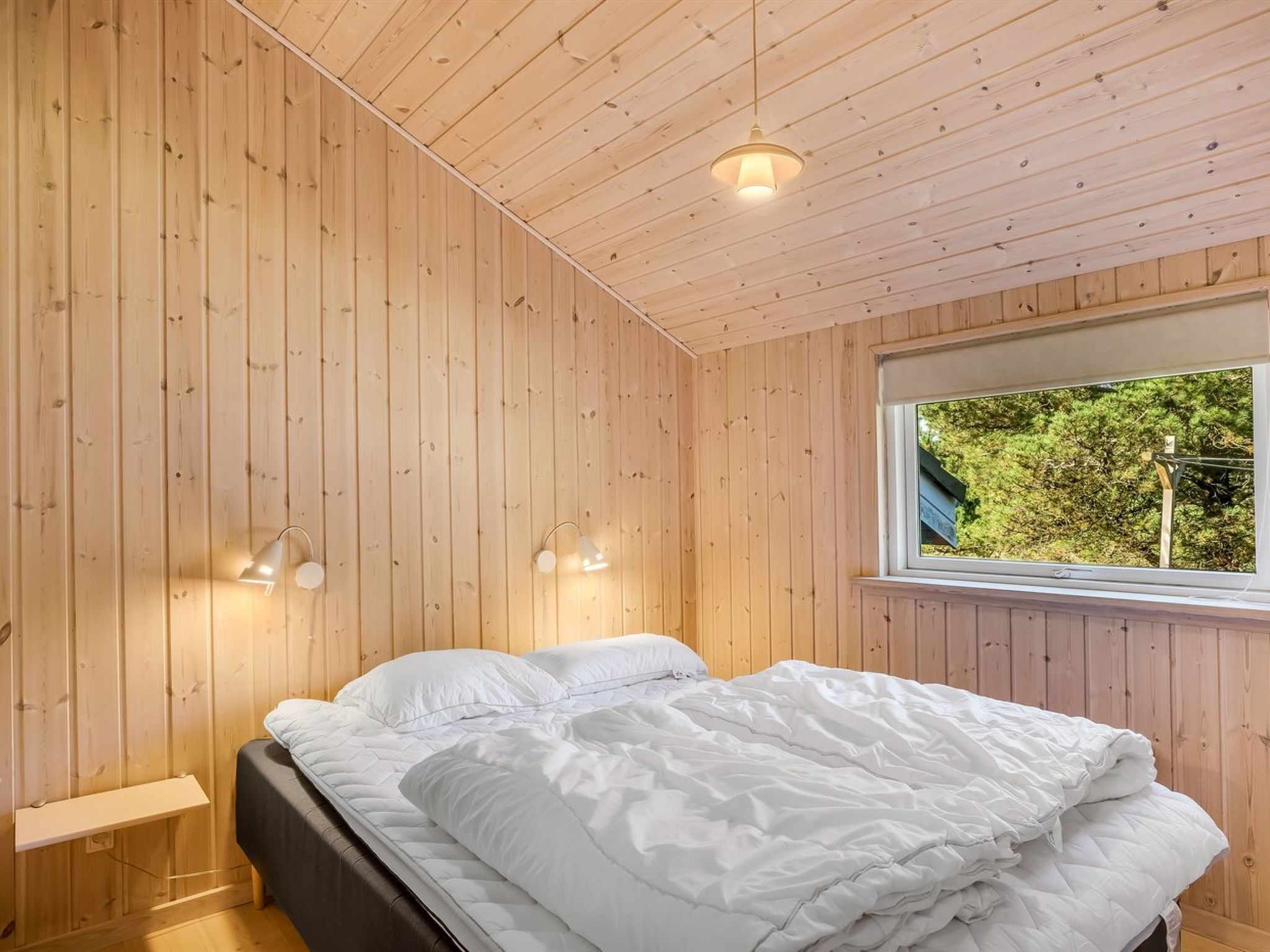 A bedroom with wooden walls, a bed with white bedding, two bedside lamps, and a window with a view of greenery.