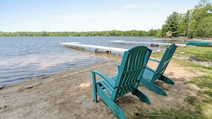 Deux chaises Adirondack vertes sont placées sur une plage de sable, au bord d’un lac paisible, avec une jetée qui s’avance dans l’eau.