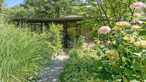 Un sentier de jardin mène à une structure couverte dotée d’un mur en pierre, le tout étant entouré d’une végétation luxuriante et de plantes en fleurs.