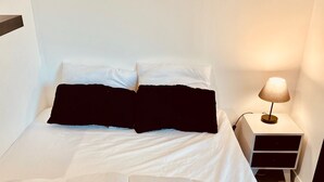 A neatly made bed with white bedding and two black pillows, a bedside table with a lamp, and a white wall. - Bright apartment, quiet, secure residence, swimming pool/parking, near Port Minimes (La Rochelle)