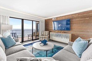 Een moderne woonkamer met een bank, fauteuil en salontafel. Er is een televisie aan de muur bevestigd en een uitzicht op de oceaan door grote ramen.