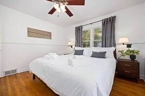 A neatly made bed with white linens and gray pillows, a wooden nightstand with a lamp, and a ceiling fan with three lights.