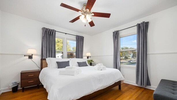 Un dormitorio con cama, lámparas de noche, mesita de noche, ventilador de techo y ventana con cortinas.