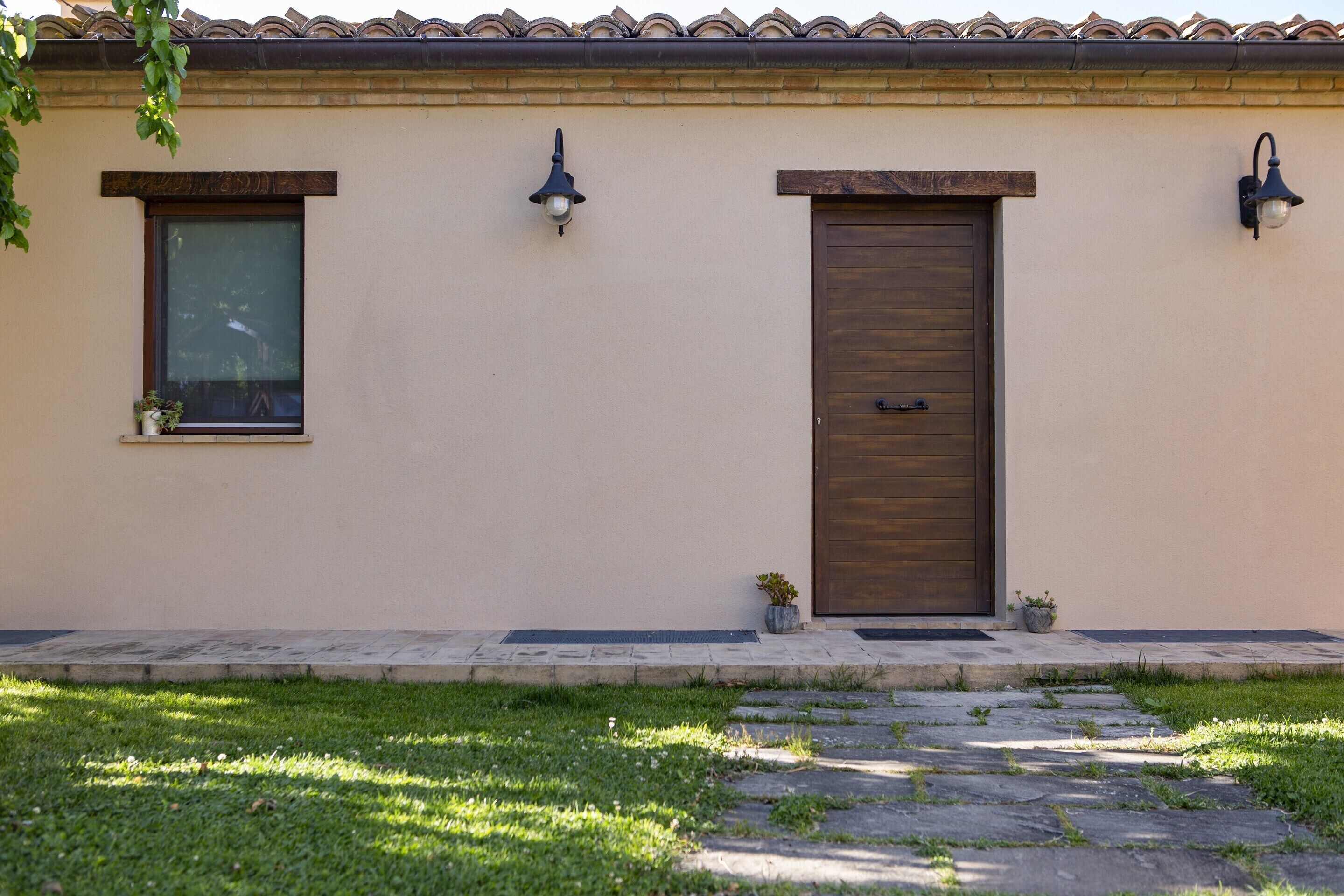 Un edificio con fachada color beige, techo de tejas, puerta de madera y dos ventanas adornadas con plantas en macetas.