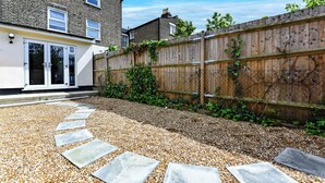 Un jardin à l’arrière d’une maison, avec un chemin de gravier, une clôture en bois et une maison en briques.