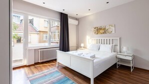 Une chambre à coucher moderne avec un lit blanc, une table de chevet, un radiateur et une fenêtre avec des rideaux.