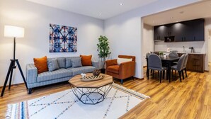 Un salon moderne comprenant un canapé, un fauteuil et une table basse, un coin repas avec des chaises, et une cuisine équipée de placards et d’un plan de travail.