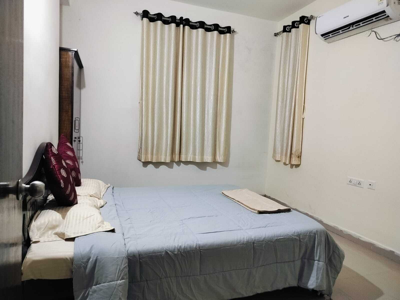 A small, single-bed room with a light blue bedspread, two beige curtains, a wooden headboard, and a wall-mounted air conditioner.