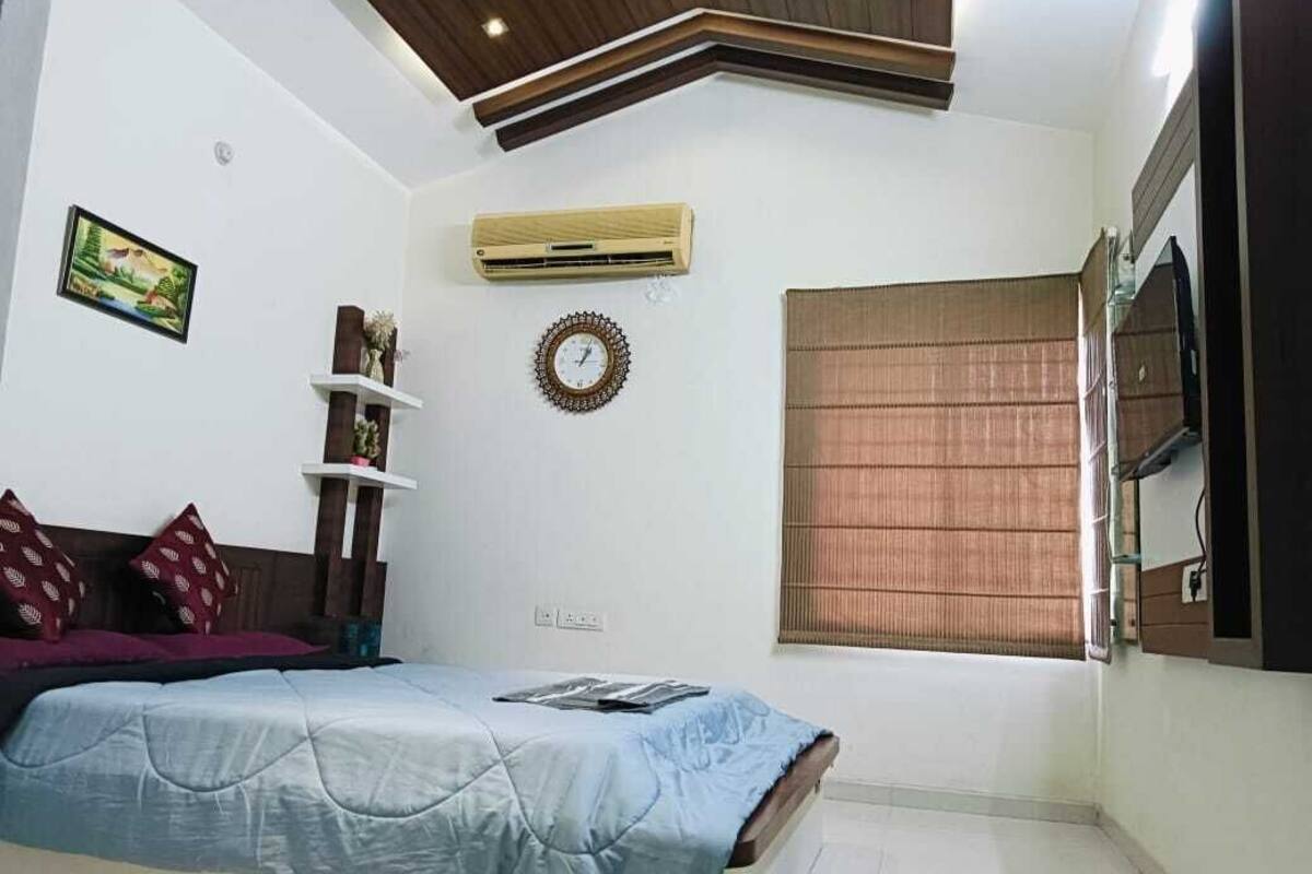 A bedroom with a bed, a wall-mounted TV, a window with blinds, a wall clock, and an air conditioning unit.