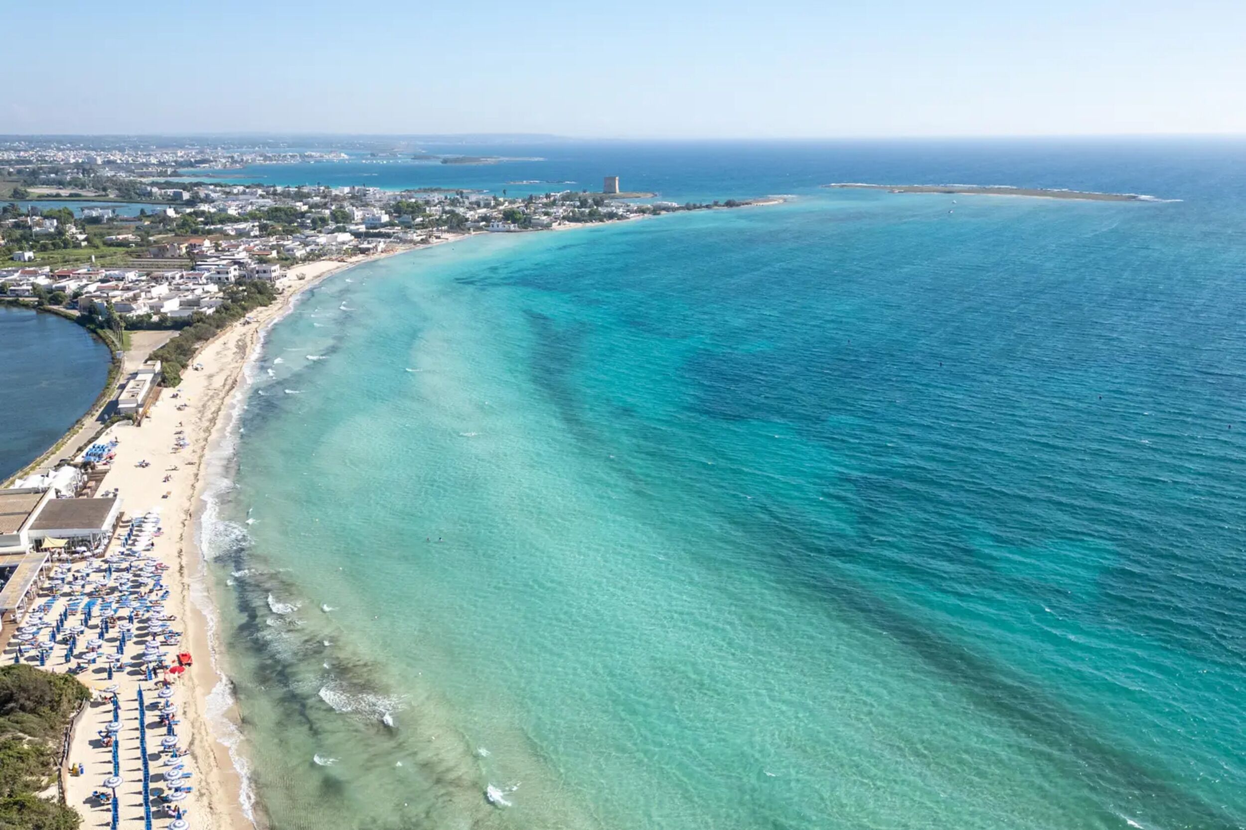 Une vue aérienne d’une zone côtière avec une plage, des eaux turquoise et limpides, et une silhouette de ville qui se dessine à l’horizon.