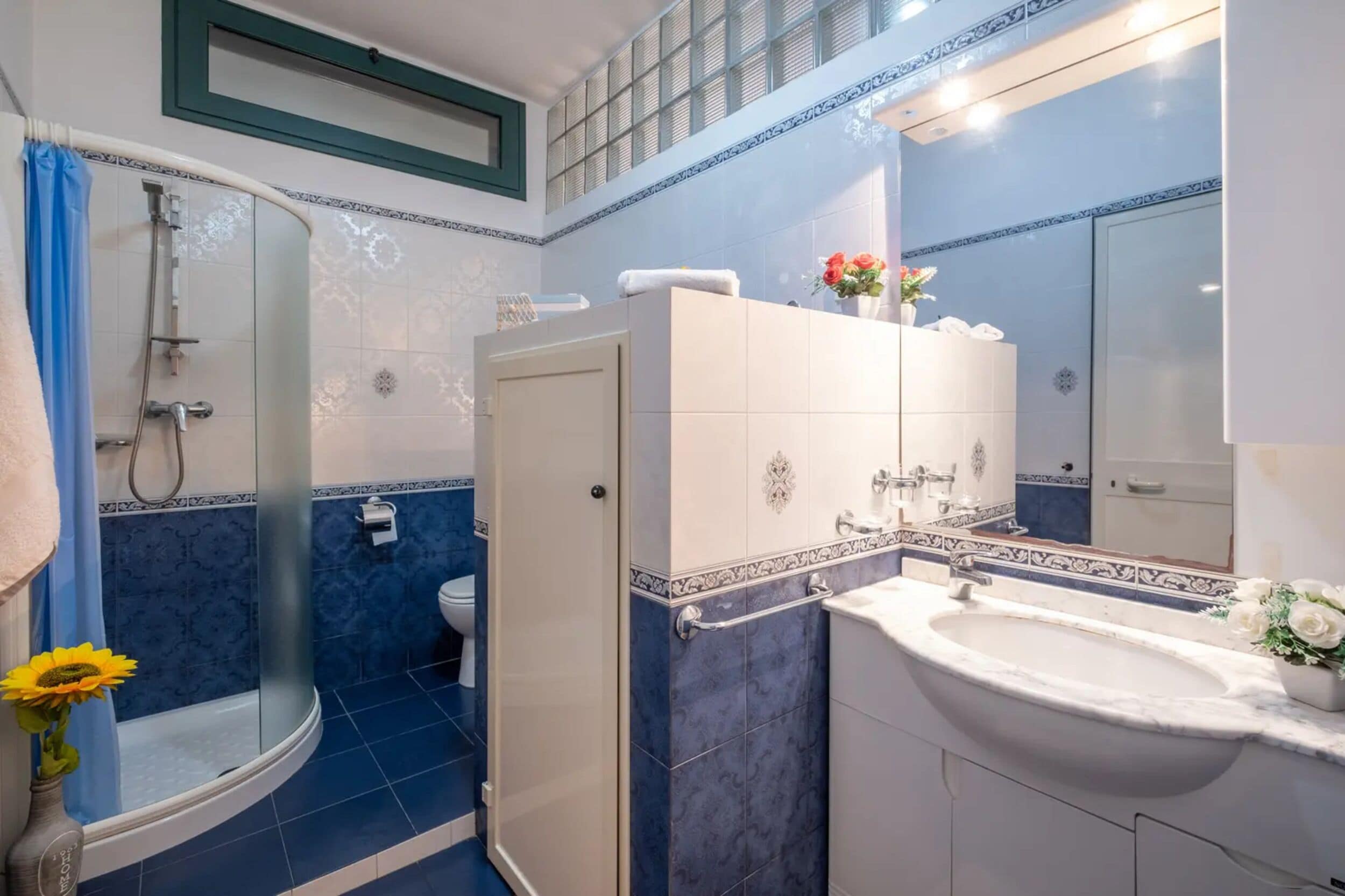 Casa de banho com chuveiro, sanita e lavatório, com azulejos azuis e brancos e um espelho.