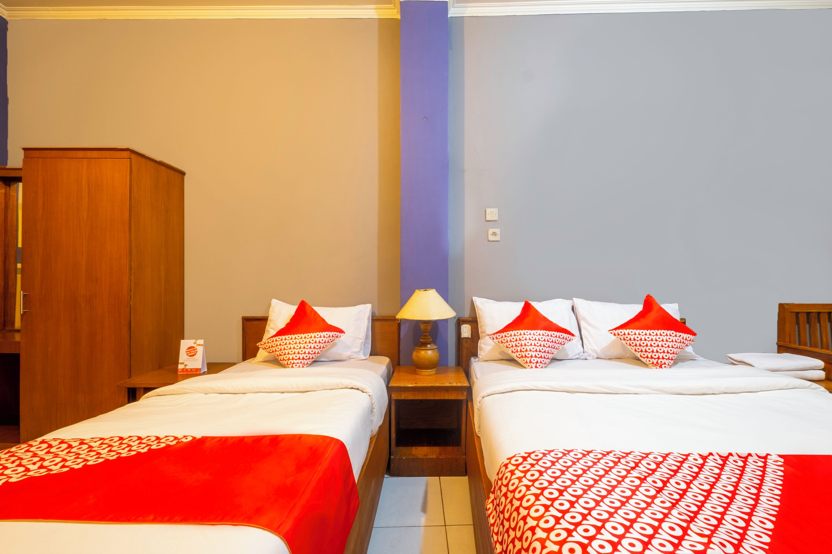 A hotel room with two beds, wooden headboards, a nightstand with a lamp, and a wardrobe.