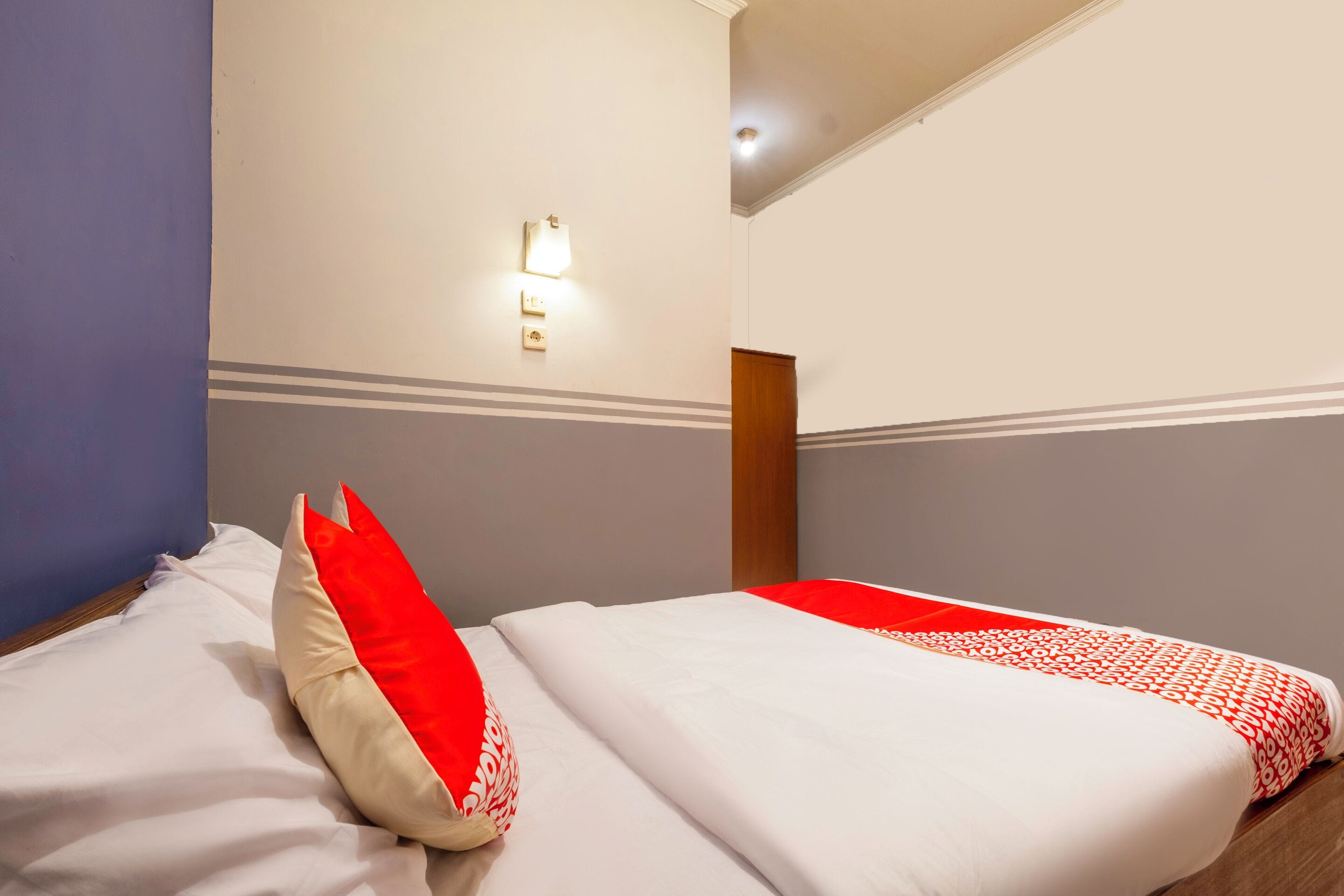 A hotel room with two beds, one with a red and white patterned bedspread, and a wall-mounted light fixture.