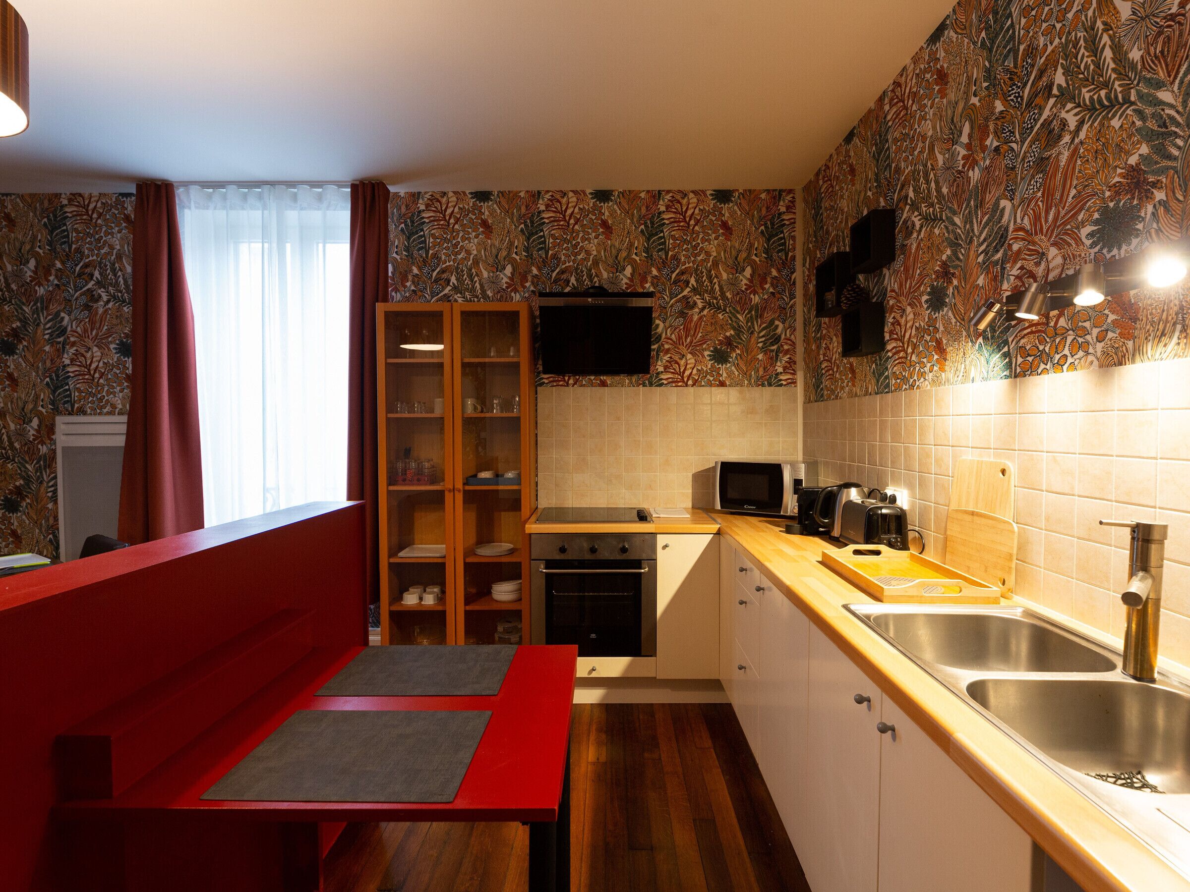 A compact kitchen with a microwave, toaster, and sink. There's a wooden cabinet with glass doors and a red bench.