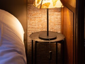 A neatly made bed with a white comforter and pillows, a small round table with a lamp featuring a butterfly pattern, and a textured brick wall in the background. - Art Nouveau Apartment Nancy, near Villa Majorelle (Nancy)