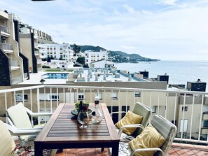 A balcony with a wooden table, white chairs, and a view of the sea and buildings.