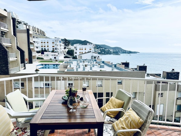 Ein Balkon mit Holztisch, weißen Stühlen und Blick auf das Meer und die Gebäude.