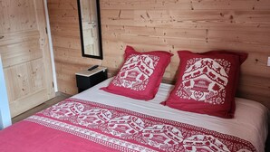 Ein Zimmer mit Holzboden, einem Bett, zwei Kissen mit rotem und weißem Muster, einem Nachttisch und einem Spiegel an der Wand.