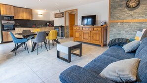 A modern living room with a sofa, coffee table, and chairs. A kitchen area with a dining table and chairs is visible in the background. - 3-Room Chalet in Châtel, 6 Pers, South Balcony (Châtel)