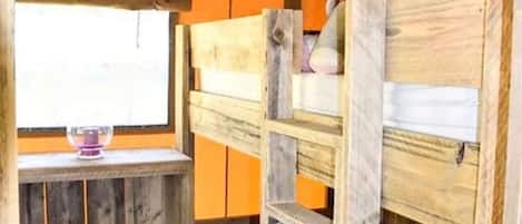 A bunk bed with a teddy bear on top, a small shelf with a candle, and a window with a view.
