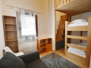 A compact room with a bunk bed, wooden shelves, a small table, and a sofa.