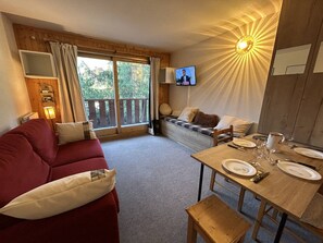 A cozy living room with a red sofa, a wooden dining table set for two, and a television mounted on the wall.