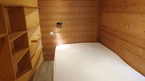 Een kleine kamer met een enkel bed, houten wanden en een schuine kap. Links bevindt zich een inbouwkast en in de muur een klein stopcontact.