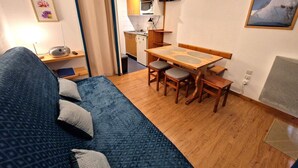 A compact living space with a sofa, a dining table with chairs, a kitchenette, a TV mounted on the wall, and a framed picture on the wall.