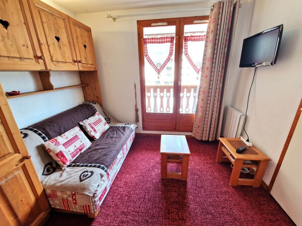 A cozy room with a wooden sofa, a small table, a TV, and a balcony with a red checkered curtain.