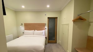 Una camera da letto con un letto, una testiera in legno, un comodino, un attaccapanni e una porta che dà su un'altra stanza.