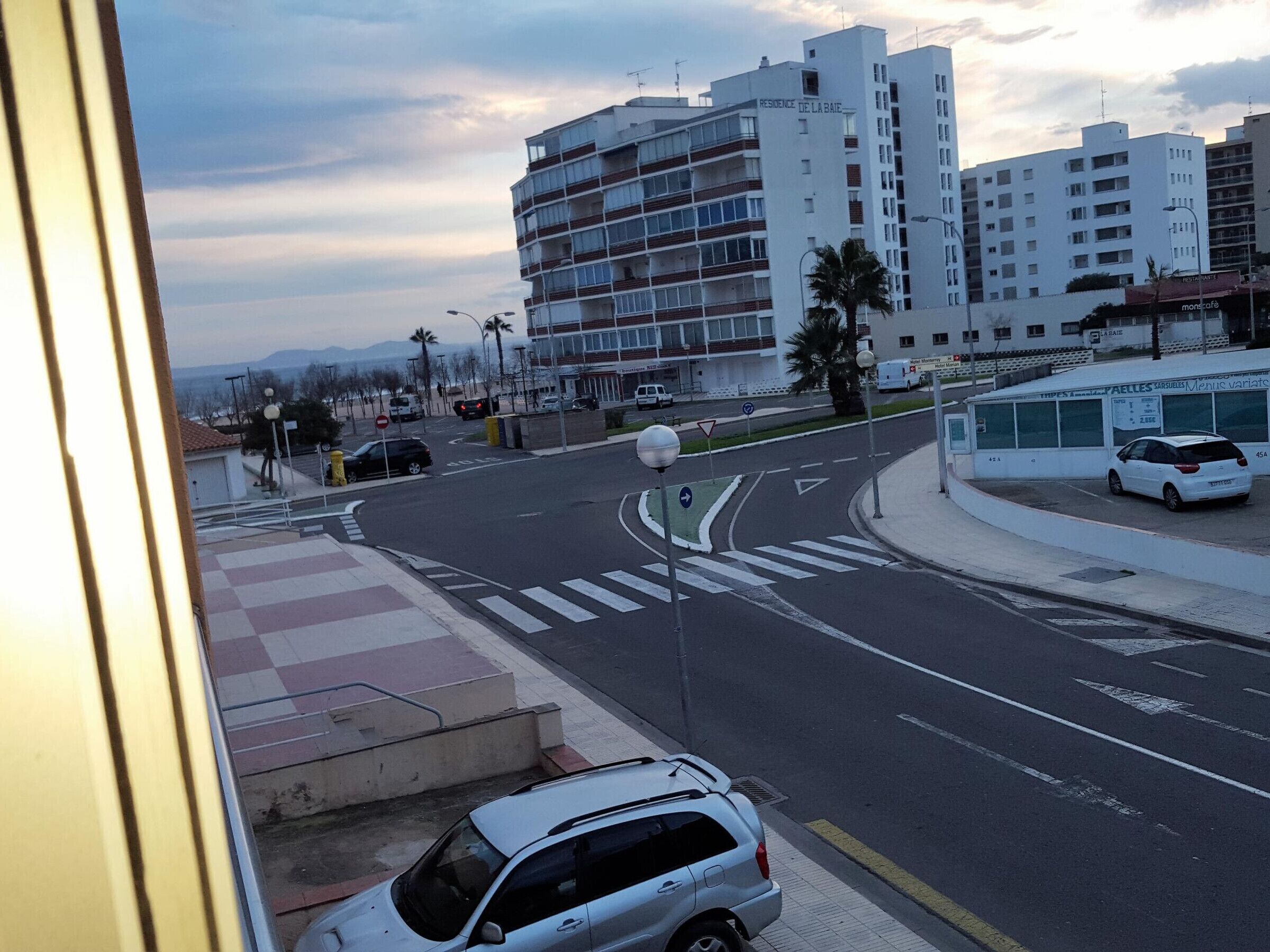 A street view with a white car parked on the side, buildings in the background, and a pedestrian crossing.