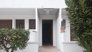 Un edificio blanco con un muro de piedra, una escalera que conduce a un acceso oscuro y plantas en macetas a la derecha.