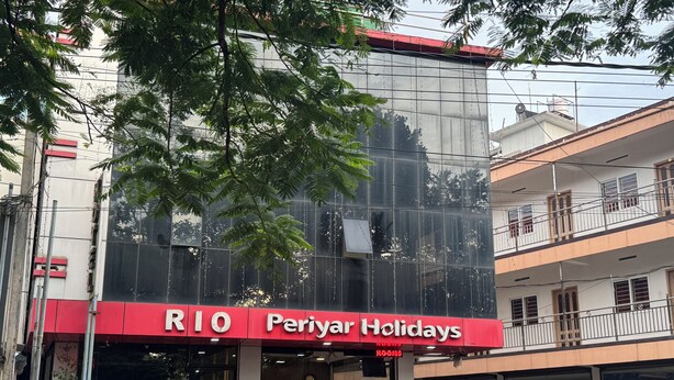 A hotel named "RIO Periyar Holidays" with a modern glass facade, a red awning, and a balcony on the upper floor. - Periyar Holidays  (Peermade)