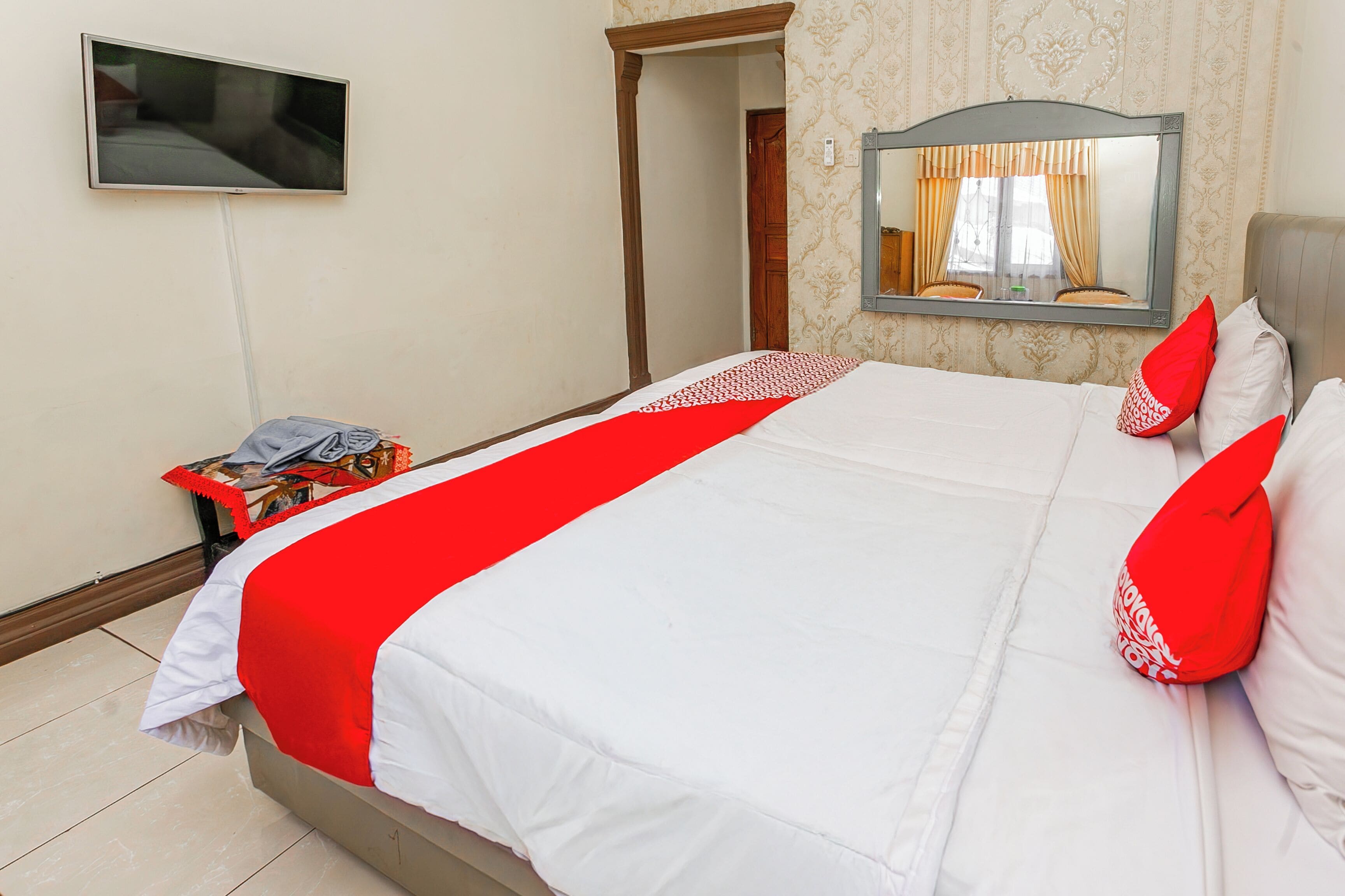 A neatly made bed with a red and white bedspread, a television mounted on the wall, and a mirror above the bed.