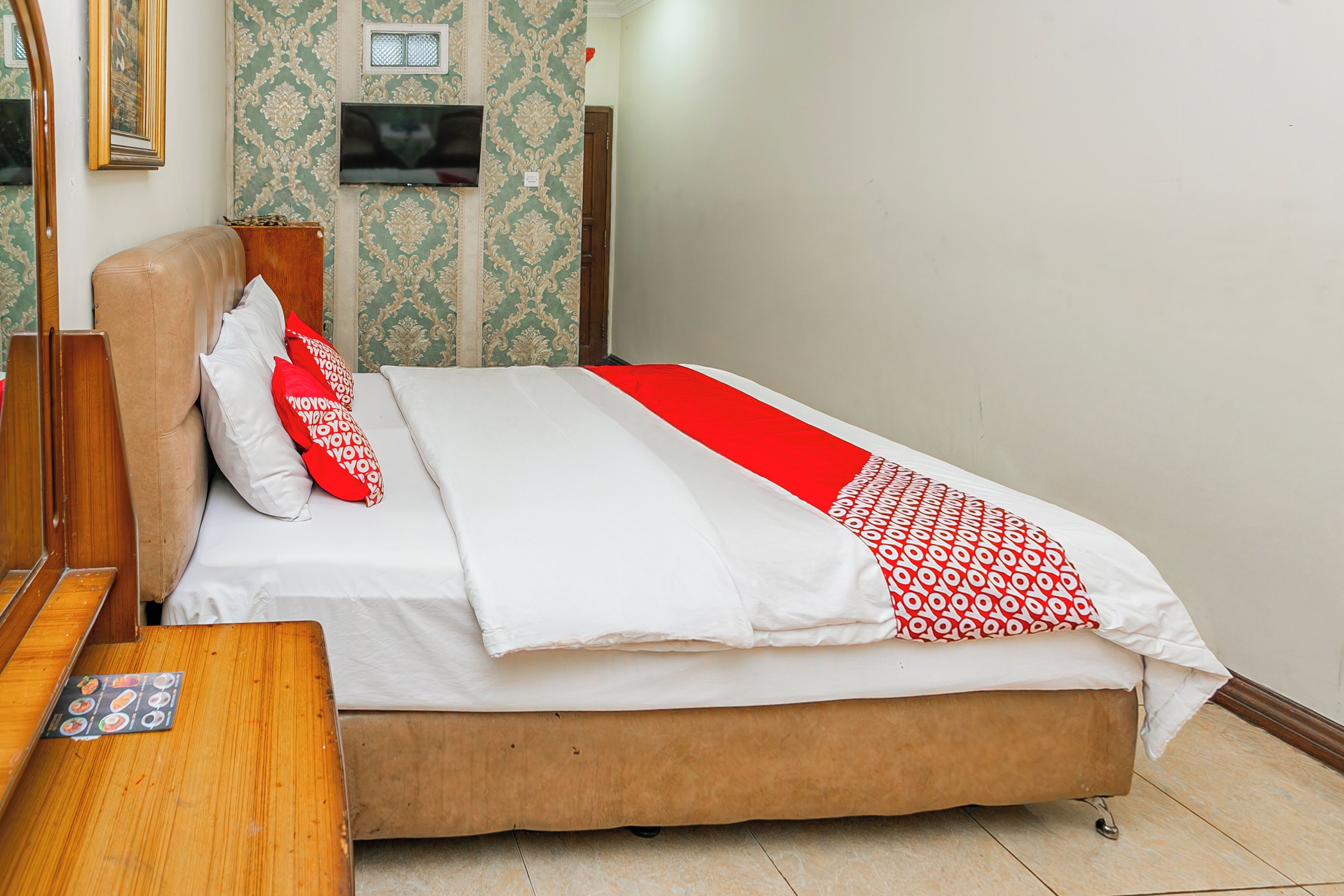 A neatly made bed with a red and white patterned blanket, a wooden headboard, and a small wooden nightstand with a menu card.