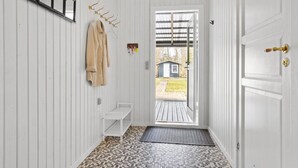 Kapea käytävä, jonka seinät ovat valkoista puuta, lattialla on kuvioitu matto, seinällä on naulakko, jossa roikkuu beige takki, ja oven läpi näkyy pieni varasto.
