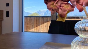 A room with a wooden table and a vase of flowers. A large window reveals a view of mountains and a wooden fence. - Attic Apartment in Saalfelden Near Ski Slopes (Saalfelden am Steinernen Meer)