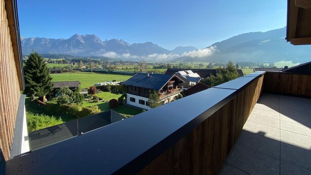 A balcony with a view of a mountainous landscape, a green field, and a building with solar panels. - Attic Apartment in Saalfelden Near Ski Slopes (Saalfelden am Steinernen Meer)