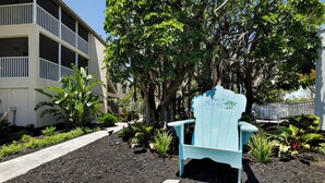 Een blauwe Adirondack-stoel met een bordje met daarop 'Sandy's Beach House' in een aangelegde tuin met mulch, planten en een gebouw op de achtergrond.