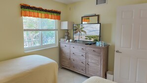 Een slaapkamer met een bed, een dressoir, een televisie en een raam met uitzicht op bomen.