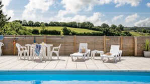 Zona junto a la piscina con sillas y mesas de plástico blanco, una valla de madera y vistas a un paisaje verde al fondo.