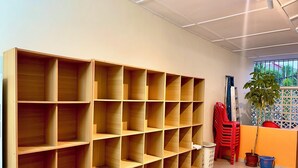 Egy tágas szoba, melyben a fal mentén egy nagy, fából készült polcrendszer áll, egy piros szék, egy virágos növény és egy fehér szemeteskosár található.