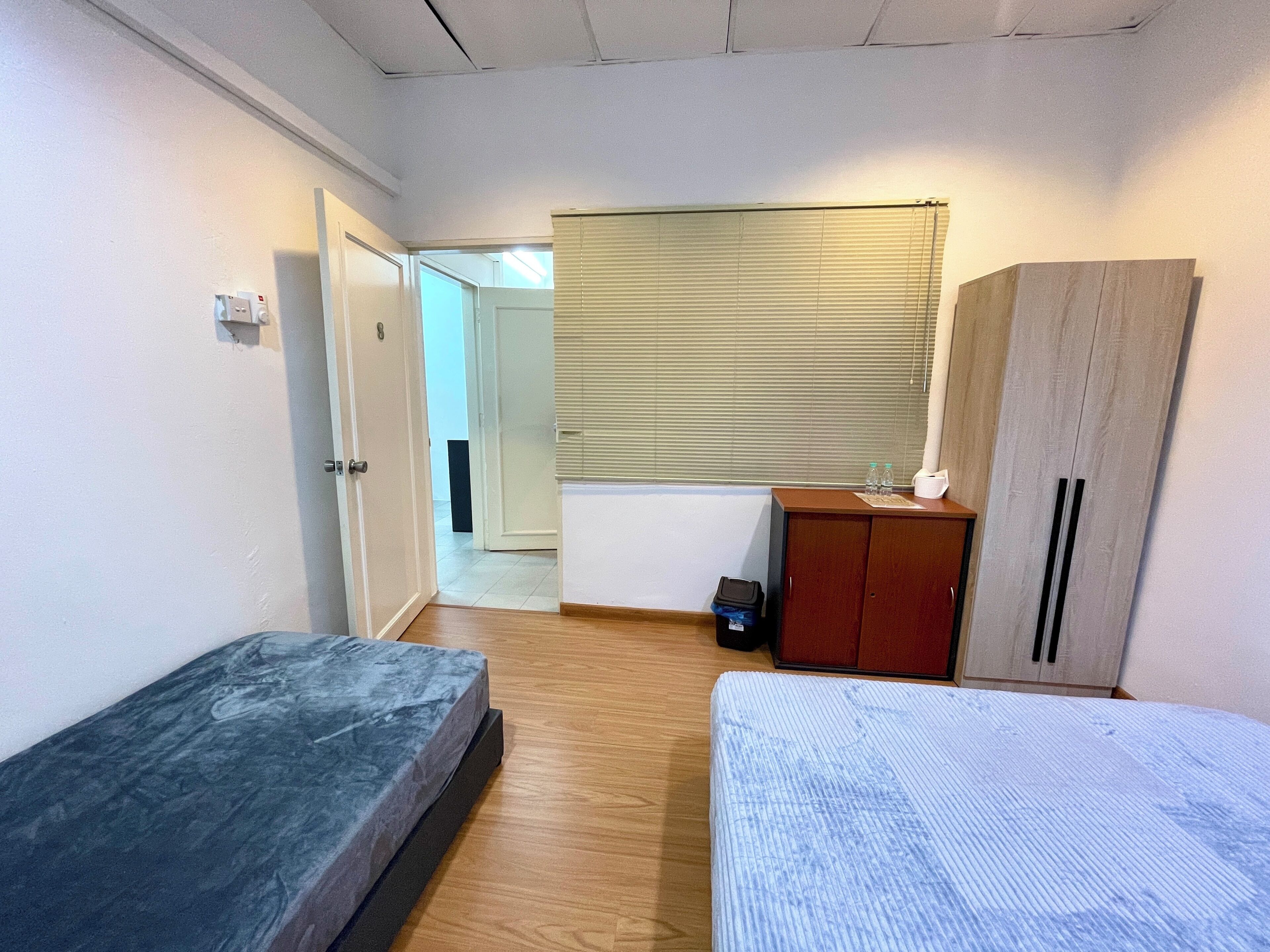 A small, sparsely furnished room with a bed, a wardrobe, a radiator, and a window with blinds.