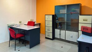 Egy szállodai szoba íróasztallal, két piros székkel, fiókos szekrénnyel és üveg vitrinnel.