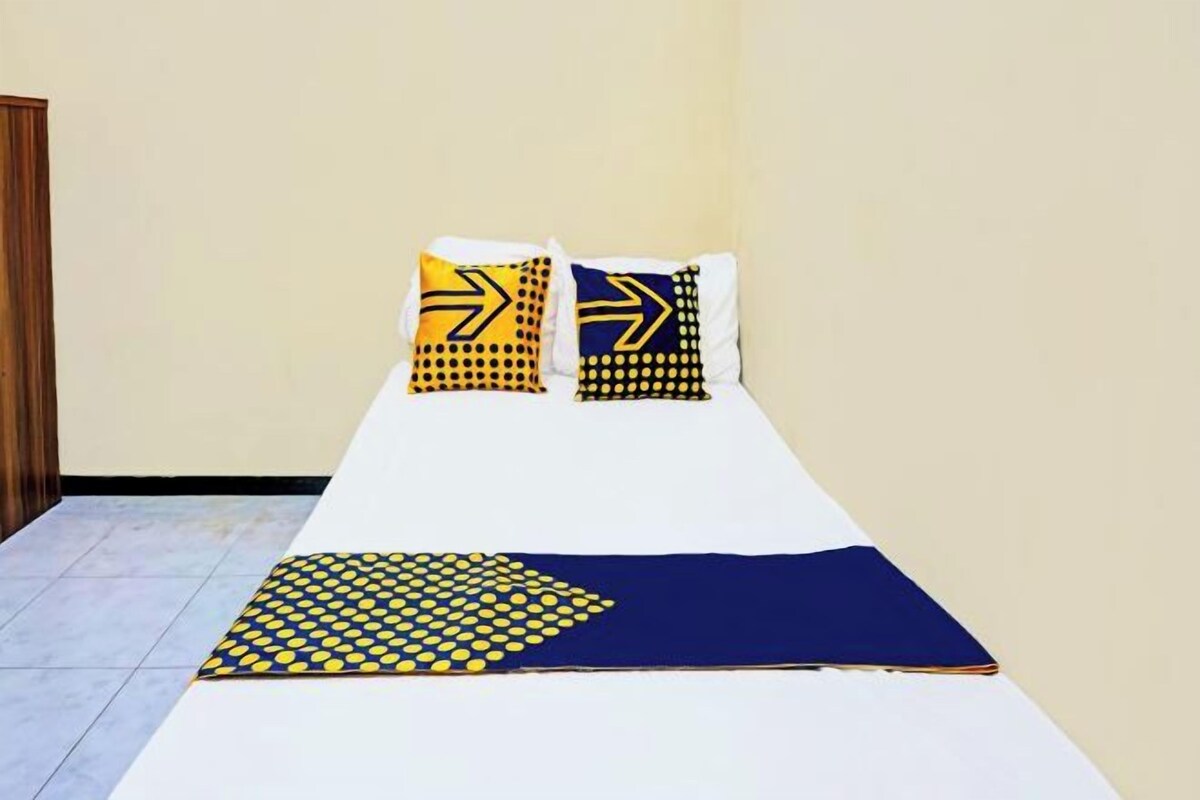 A single bed with a white sheet, two pillows with yellow and black patterns, and a blue blanket with yellow dots.
