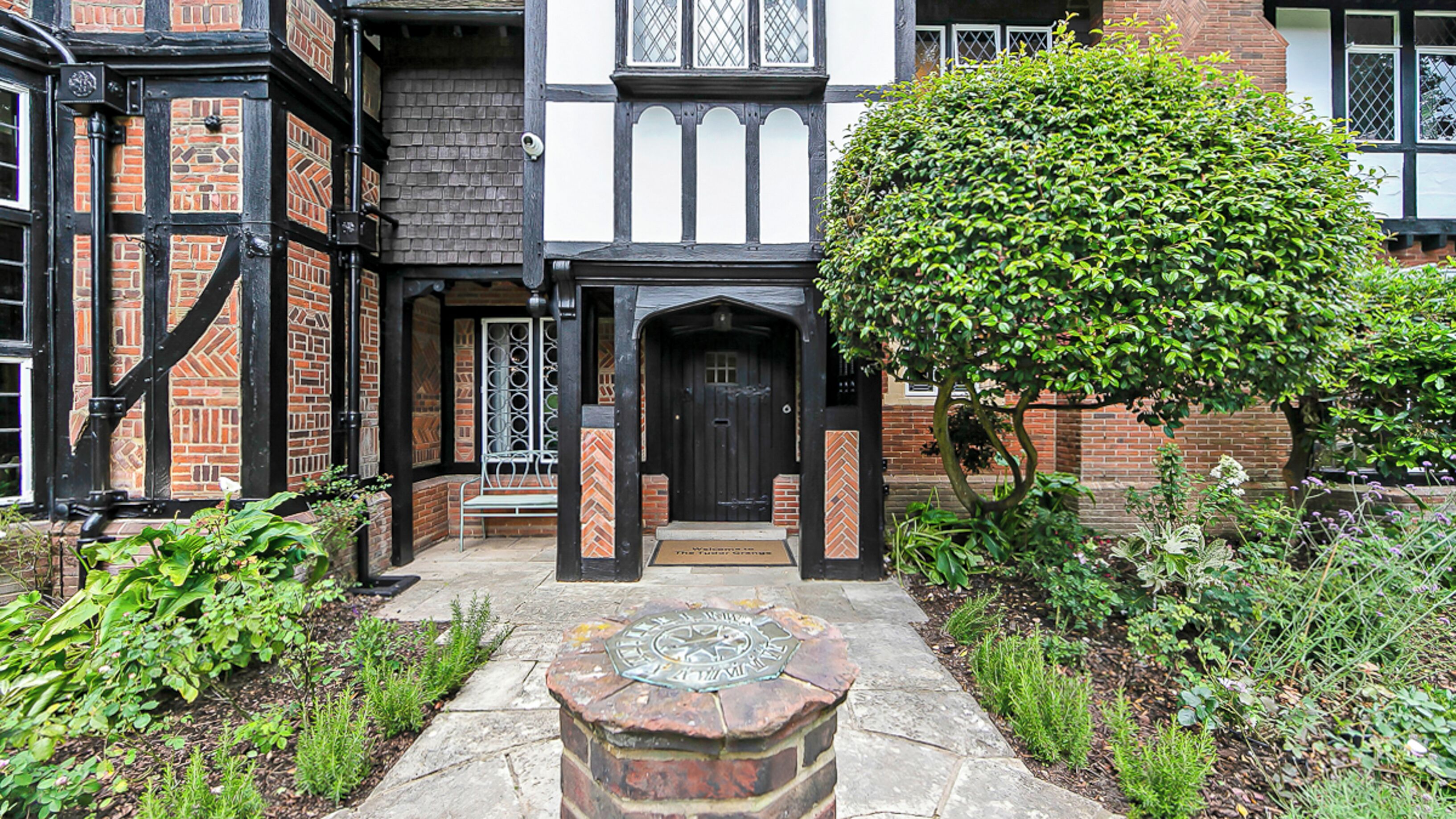A traditional half-timbered house with a black door, a small garden with a stone bench, and a large tree.