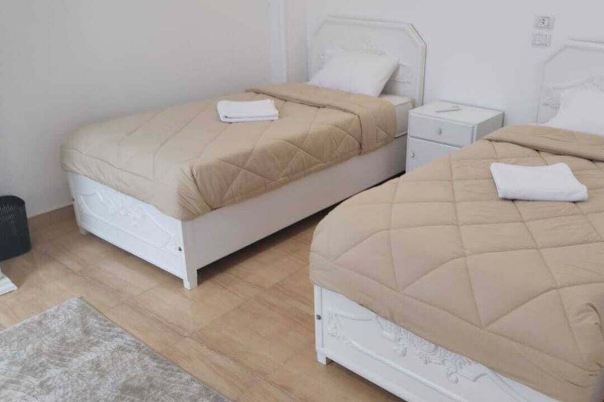 Two twin beds with beige bedding and white headboards in a room with light wooden flooring.