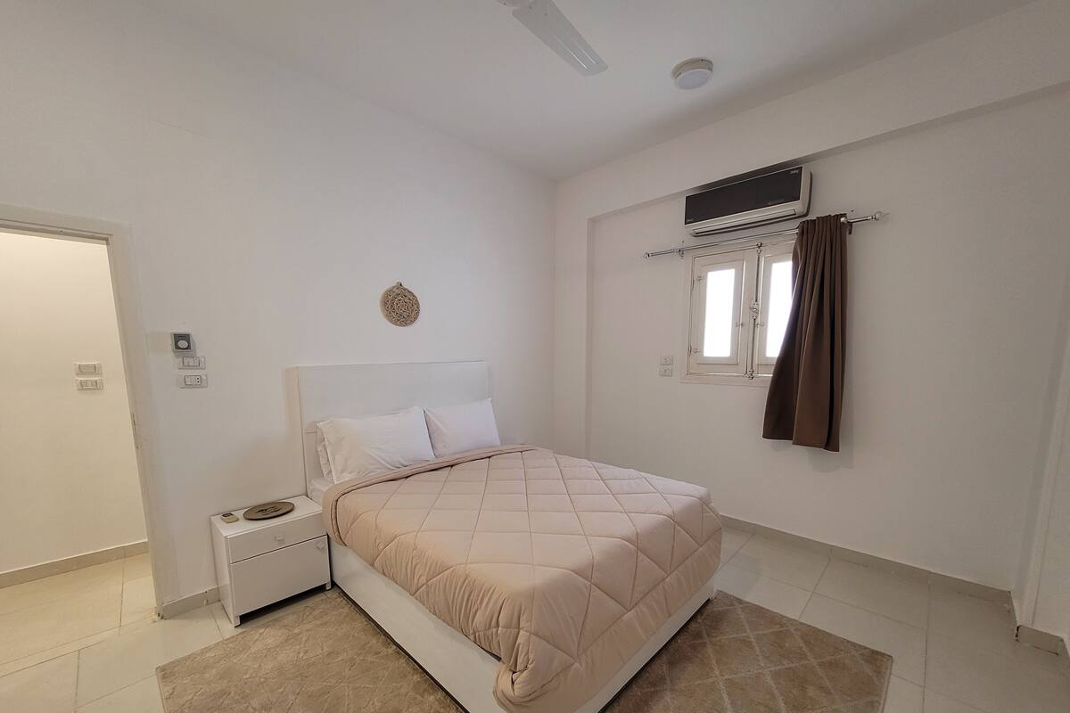 A bedroom with a bed, a nightstand, a wall-mounted air conditioner, and a window with curtains.