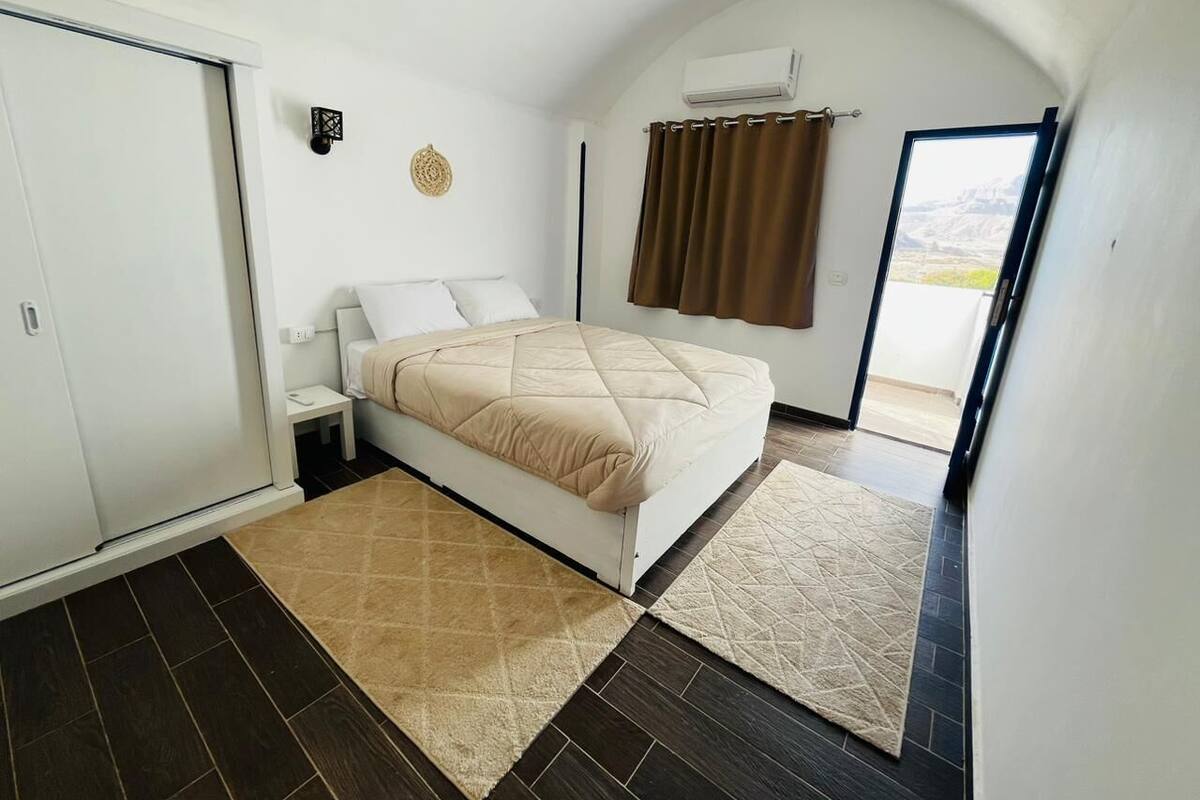A bedroom with a bed, a nightstand, a wall-mounted air conditioner, and a view through a door.