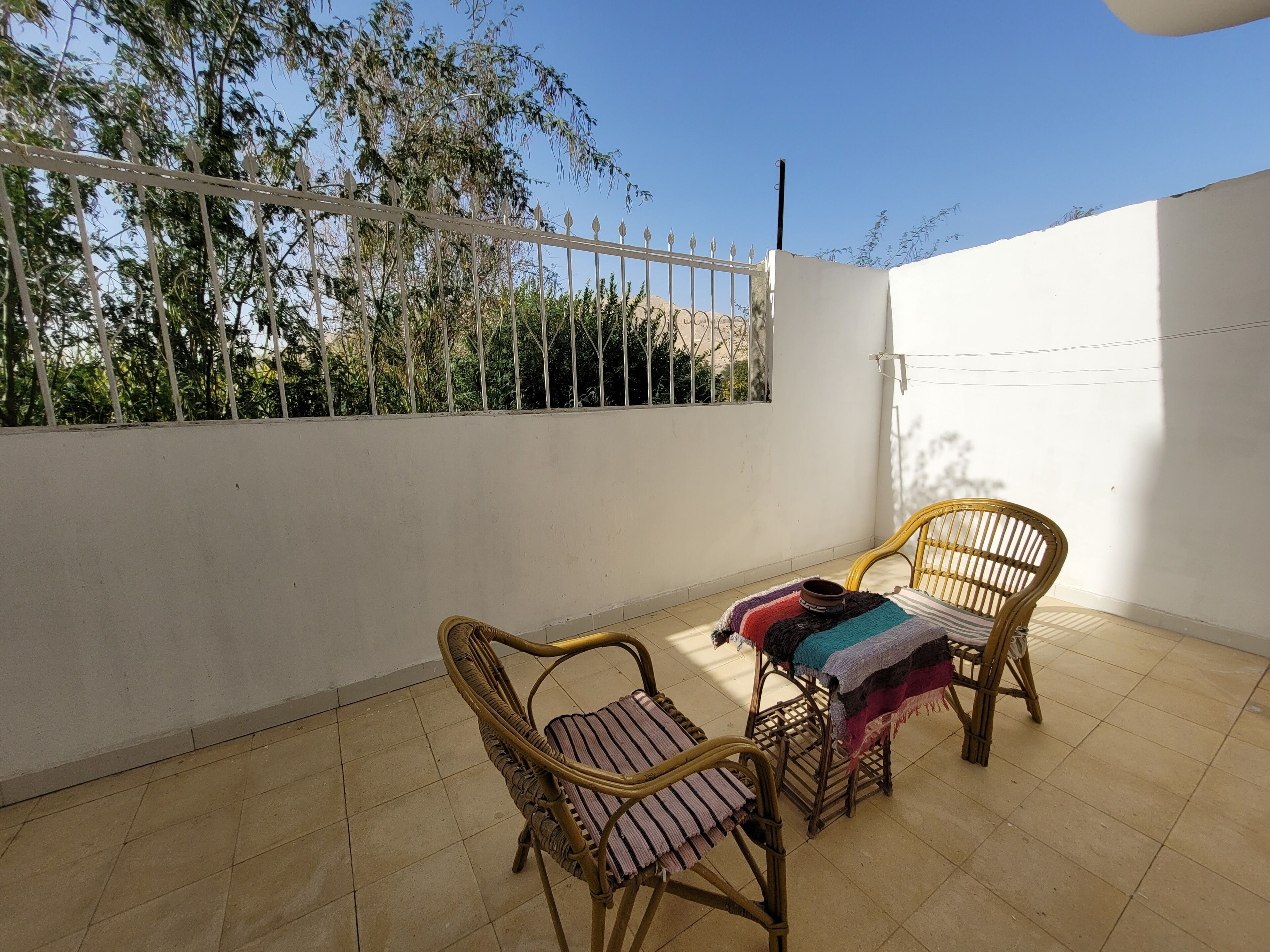 Un balcon avec deux fauteuils en osier et une petite table, un mur blanc et un portail en métal.